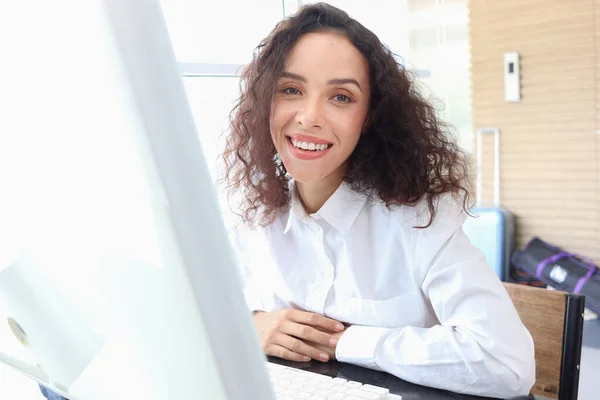 Portrait of happy smiling young beautiful woman with curly hair using desktop computer, female officer staff in casual white shirt sitting at working desk at office workplace.