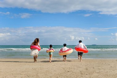 Ebeveynleri renkli yüzme yüzüğü takan çocuklar mavi denize koşuyor, çocuklar ve yetişkinler tropikal kumsalda kuyrukta, aileleri yaz tatilinde birlikte vakit geçiriyor.. 