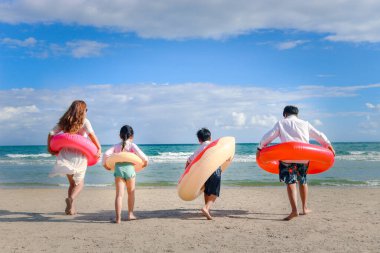 Ebeveynleri renkli yüzme yüzüğü takan çocuklar mavi denize koşuyor, çocuklar ve yetişkinler tropikal kumsalda kuyrukta, aileleri yaz tatilinde birlikte vakit geçiriyor.. 