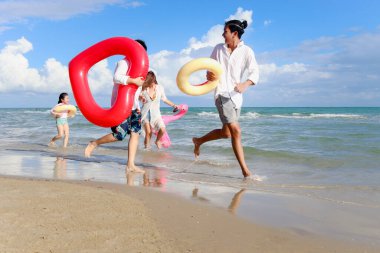 Ebeveynleri renkli yüzme yüzüğü takan çocuklar yaz plajlarında koşuşturuyor, çocuklar ve yetişkinler tropik kumsalda, mutlu bir aile olarak vakit geçiriyorlar ve yaz tatilinde birlikte eğleniyorlar.. 