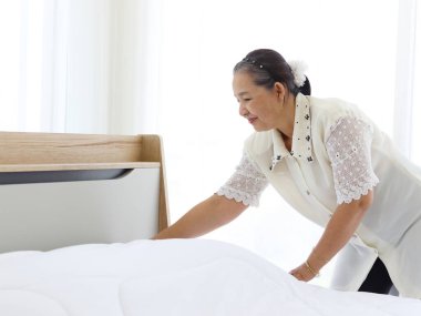 Mutlu gülümseyen Asyalı yaşlı ev hanımı yatak odasını temizliyor, büyükannem ev işi yapıyor ve temizlik yapıyor, yaşlı kadın oda hizmetçisi beyaz battaniye ve çarşafı değiştiriyor..