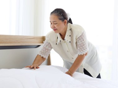 Mutlu gülümseyen Asyalı yaşlı ev hanımı yatak odasını temizliyor, büyükannem ev işi yapıyor ve temizlik yapıyor, yaşlı kadın oda hizmetçisi beyaz battaniye ve çarşafı değiştiriyor.