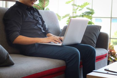 Asyalı genç işadamı evinde kanepesinde otururken klavye bilgisayarında daktilo kullanıyor. Oturma odasında online çalışan serbest çalışan biri.. 