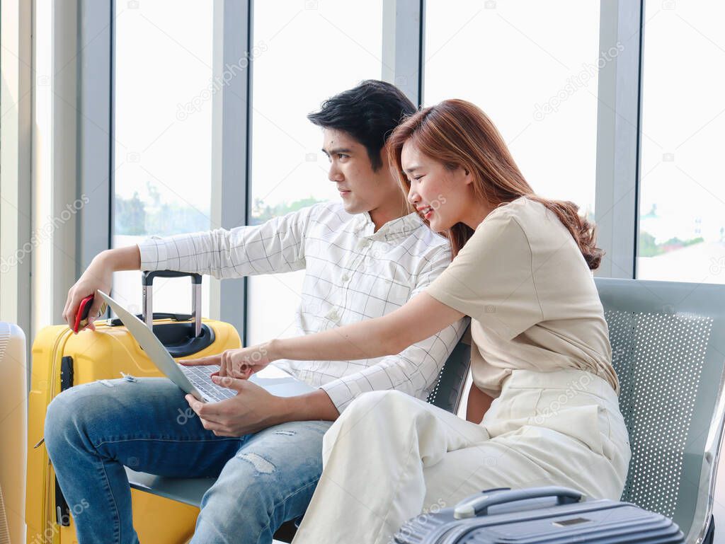 Asian young traveler lover couple siting on terminal chair seat with luggage suitcase, using laptop computer together, talking having and having fun times, happy friend traveling on vacation. 