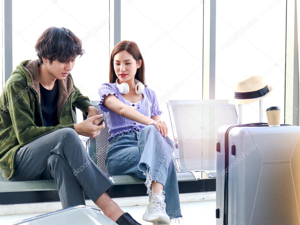 Asian young traveler lover couple siting on terminal chair seat with luggage suitcase, having conversation, laughing and having fun times together, happy friend traveling on vacation. 