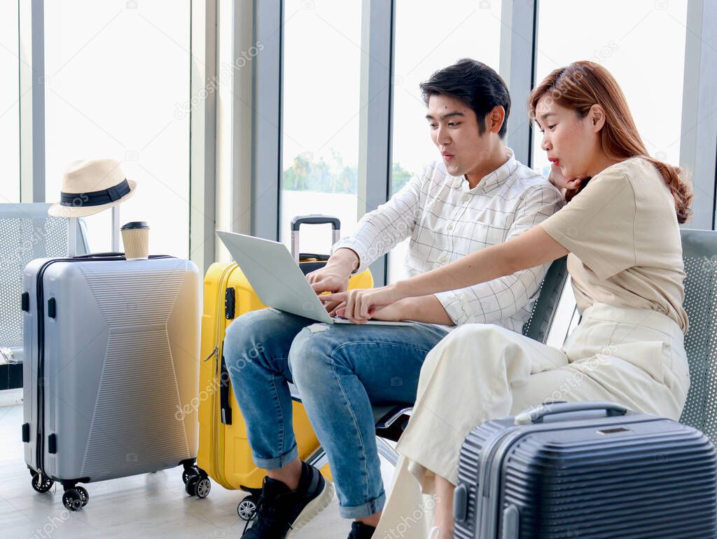 Asian young traveler lover couple siting on terminal chair seat with luggage suitcase, using laptop computer together, talking having and having fun times, happy friend traveling on vacation. 