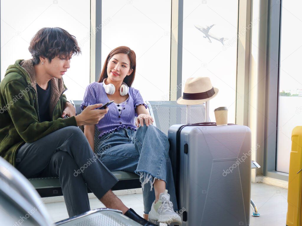 Asian young traveler lover couple siting on terminal chair seat with luggage suitcase, having conversation, laughing and having fun times together, happy friend traveling on vacation. 