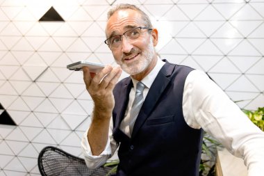 Modern ofisteki akıllı telefondan mutlu kıdemli iş adamları konuşuyor. Yaşlı adam iş yerinde yapay zeka teknolojisiyle bağlantı kuruyor. Olgun yönetici uzaktan kontrol etmek için cep telefonu kullanır.