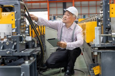 Asyalı usta ustabaşı elinde tabletle fabrikada makineleri incelerken. Sanayi bölgesinde makineyle çalışırken tablet kullanan miğferi olan yaşlı bir yönetici.