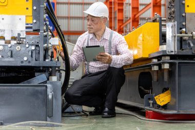 Asyalı usta ustabaşı elinde tabletle fabrikada makineleri incelerken. Sanayi bölgesinde makineyle çalışırken tablet kullanan miğferi olan yaşlı bir yönetici.