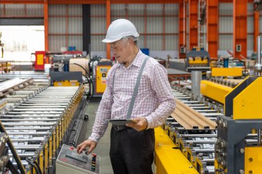 Asyalı usta ustabaşı elinde tabletle fabrikada makineleri incelerken. Sanayi bölgesinde makineyle çalışırken tablet kullanan miğferi olan yaşlı bir yönetici.