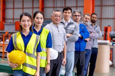Bir grup mutlu mühendis imalat fabrikasında sıraya dizildi. Mühendislerin miğferi vardır, sanayi bölgesinde güvenle dururlar. Çeşitli profesyonel takımlar kameraya vizyonlarla bakar..