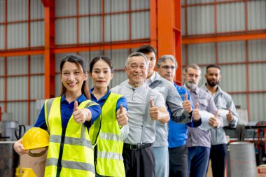 Bir grup mutlu mühendis imalat fabrikasında sıraya dizildi. Kurumsal mühendislik işçileri miğfer tutarlar, endüstriyel alanda güvenle başparmaklarını kaldırırlar. Çeşitli takımlar kameraya görüntülerle bakar.