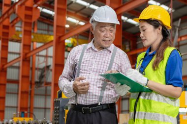 Miğfer ve güvenlik yeleği olan Asyalı kadın işçi pano tutuyor, endüstri alanındaki yöneticiyle tartışıyor. Ustabaşı üretim fabrikasında inceleme yaparken personele talimat veriyor..