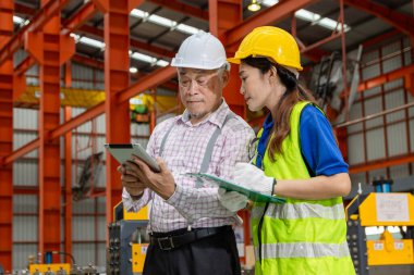 Miğfer ve güvenlik yeleği olan Asyalı kadın işçi pano tutuyor, endüstri alanındaki yöneticiyle tartışıyor. Ustabaşı üretim fabrikasında inceleme yaparken personele talimat veriyor..