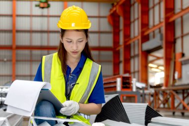 Miğfer ve güvenlik yeleği olan Asyalı kadın bir pano tutuyor ve imalat fabrikasında çalışıyor. Kadın mühendis, endüstriyel alandaki metal levhaları inceliyor. Müfettiş ürünün kalitesini kontrol ediyor..