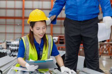Miğfer ve güvenlik yeleği olan Asyalı kadın işçi pano tutuyor, endüstri alanındaki yöneticiyle tartışıyor. Üretim fabrikasında makineleri denetlerken ustabaşı personele talimat verir.