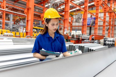 Güvenlik kaskı takan Asyalı kadın bir pano tutuyor, imalat fabrikasında çalışıyor. Kadın mühendis, endüstriyel alandaki metal levhaları inceliyor. Müfettiş ürünün kalitesini kontrol ediyor..