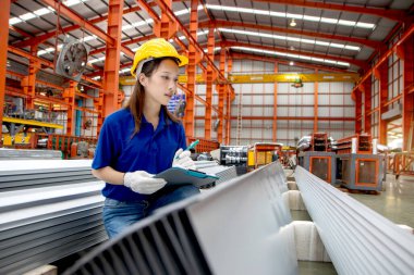 Güvenlik kaskı takan Asyalı kadın bir pano tutuyor, imalat fabrikasında çalışıyor. Kadın mühendis, endüstriyel alandaki metal levhaları inceliyor. Müfettiş ürünün kalitesini kontrol ediyor..