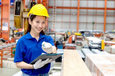 Güvenlik kaskı takan Asyalı kadın bir pano tutuyor, imalat fabrikasında çalışıyor. Kadın mühendis, endüstriyel alandaki metal levhaları inceliyor. Müfettiş ürünün kalitesini kontrol ediyor..