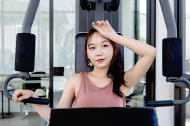 Spor aletleriyle spor yapan Asyalı güzel bir kadın. Göğüs presi makinesi kullanırken alnındaki teri silen fitness kızı. Sportif kadın vücut geliştirme antrenmanı yapıyor..