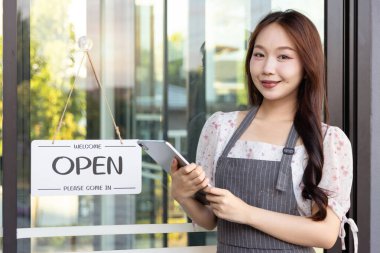 Önlüklü Asyalı güzel bir kadın tableti tutuyor, kafenin camlı kapısında açık tabelanın yanında duruyor. Küçük bir işletme kuran genç sahibi açık tabela. Kahve dükkanında çalışan mutlu bayan garson..