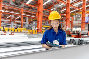 Güvenlik kaskı takan Asyalı kadın bir pano tutuyor, imalat fabrikasında çalışıyor. Kadın mühendis, endüstriyel alandaki metal levhaları inceliyor. Müfettiş ürünün kalitesini kontrol ediyor..