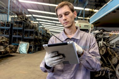 Fabrikadaki motor parçalarını incelerken telsiz ve tablet kullanan güvenlik koruyucu üniformalı genç mühendis. Yedek parça deposunda çalışan endüstriyel işçiler..