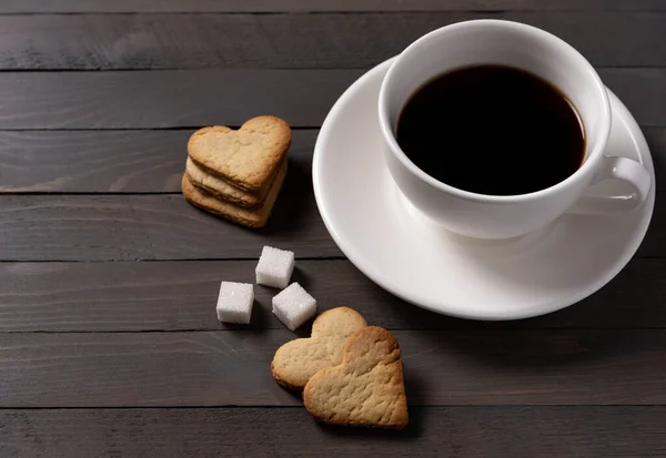 black coffee in a white cup on a white saucer with heart-shaped cookies and sugar cubes on a brown wooden background horizontal photo side view