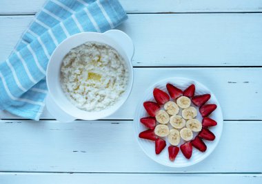 Beyaz bir seramik tencerede bir dilim eritilmiş tereyağlı sütlü pirinç lapası beyaz ahşap arka planda beyaz bir tabakta çilek ve muz dilimlerinin yanında mutfak havlusu ile birlikte.