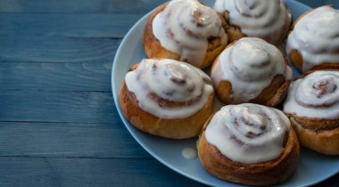 Mavi ahşap arka planda, sağdaki mavi tabakta beyaz kremayla kaplı ev yapımı, gül rengi çörekler.