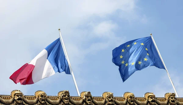 The national flag of France and the flag of the European Union against a blue sky. Flags of the European Union and the French Republic on the pediment of the Grand Opera with Golden theatrical masks. Paris