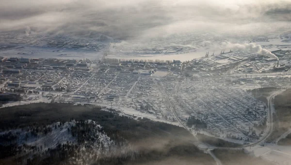 The city of Ust-Kut on the Lena river in winter in a frosty fog and haze, photo from the plane. Snow-covered Siberian taiga and Osetrovo microdistrict among the hills in December through the plane window. Irkutsk region