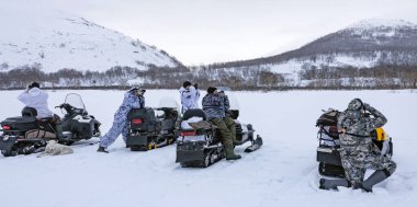 Kamuflajlı kış kıyafetleri giyen adamlar etraftaki dağları dürbünle kar araçlarından inceliyorlar. Karla kaplı bir bölgede beş avcı hayvan arıyor..  