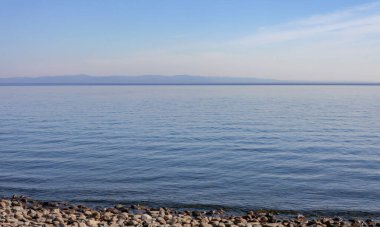 Baykal 'ın yaz sabahı çakıl taşı plajından sakin göl manzarası. Su yüzeyi ve Baykal Gölü dağlarıyla Haziran manzarası. 