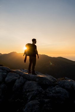 Bir dağın tepesindeki yürüyüşçünün dikey görüntüsü. Yoğun renklerle ve mavi gökyüzüyle birleşmiş, büyük bir sırt çantası taşıyan güzel altın saat günbatımının tadını çıkarıyor.