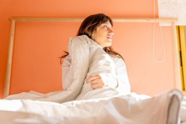 Yatakta oturup pencereye bakan genç bir kadın profili, beyaz giyen bir kadın.