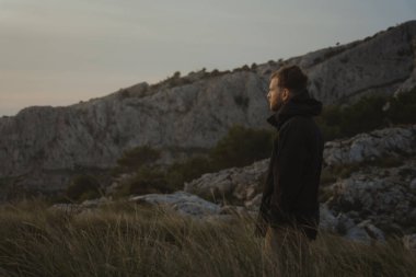 Sarışın adam güneşin doğuşunu seyrediyor. Etrafında yabani otlar olan bir dağdan altın saati bekliyor.