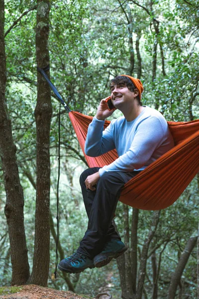 Hammock on trees in the forest ,close-up of man using mobile on a hammock in the forest, wears teal sweater and orange hat