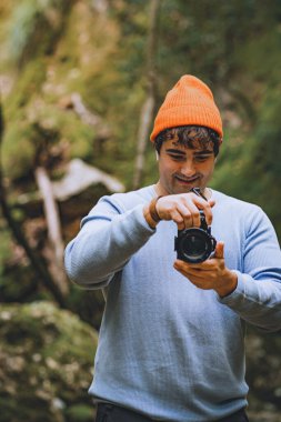 Ormanın ortasında fotoğraf makinesi olan genç adam fotoğrafçının fotoğrafını çekiyor, adam turuncu şapka ve deniz mavisi süveter giyiyor.