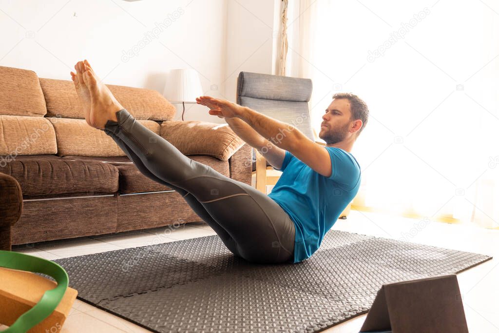 young man doing crunches at home on sports mat with raised legs, bright room with tablet and yoga blocks around.