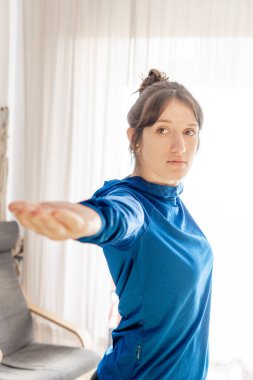 Parlak oturma odasında yoga yapan güzel bir kızın portresi, kollarını uzatmış mavi tişört giymiş bir kadın.