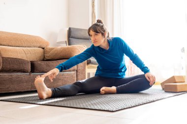 Spor kızı oturma odasında esneme egzersizleri yapıyor. Doğal ışık arka planı, spor anlayışı, yoga ve evde esneme..