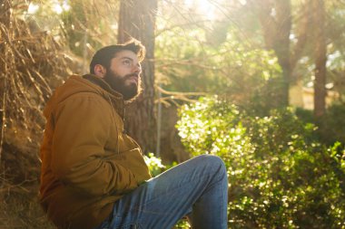 Genç adam oturuyor ve Akdeniz 'de bir manzara ormanındaki ağaçlara bakıyor. Arka planda güneş ve çok parlak bir ışık var.