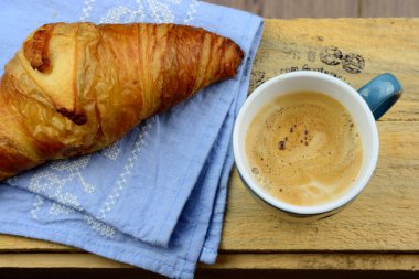 Blue coffe mug with fresh coffee and cream and croissant bread with a light grey blue tea towel with broidery on a old used wooden brakfast table in vintage style