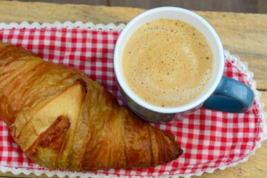 Blue coffee mug with fresh coffee and cream and croissant bread with a red white doily towel with broidery on a old used wooden breakfast table in vintage style.Good morning. Coffee time