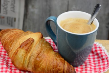 Blue coffe mug with fresh coffee and cream and croissant bread with a light grey blue tea towel with broidery on a old used wooden brakfast table in vintage style
