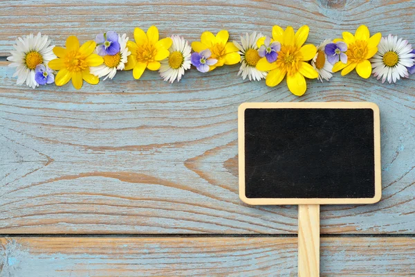 Leere schwarze Kreidetafel auf einem alten gebrauchten geknoteten blauen, grauen Holz mit Gänseblümchen und Ranunkeln Frühling und Sommer für eine heitere fröhliche Atmosphäre Blumen — Stockfoto