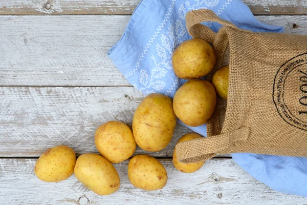 Patatas orgánicas frescas sin cocer en una bolsa de yute sobre un viejo fondo de estantes blancos de madera envejecida con toalla de cocina azul vintage con espacio de copia vacío —  Fotos de Stock