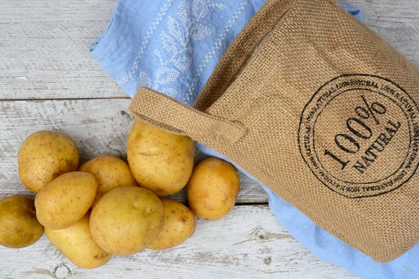 Organic fresh uncooked potatoes in a jute bag on a old weathered wooden white shelves background with vintage blue kitchen tea towel with empty copy space — Stock Photo, Image
